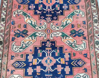 Op de afbeelding: Een rechthoekig, vintage tapijt met een centraal geometrisch ontwerp in marineblauw, roze en groen. Het tapijt heeft een herhalend patroon van gestileerde vormen en randen met een vervaagde, antieke uitstraling. De afmetingen van het tapijt zijn niet zichtbaar.