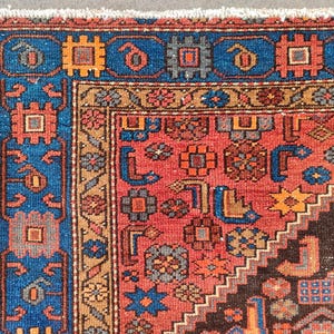 May include: Close-up of a section of a Persian rug with a detailed geometric pattern. The rug features a vibrant color palette of red, blue, orange, and brown. The border is a deep blue with repeating geometric shapes. The central field is red with intricate floral and geometric designs.