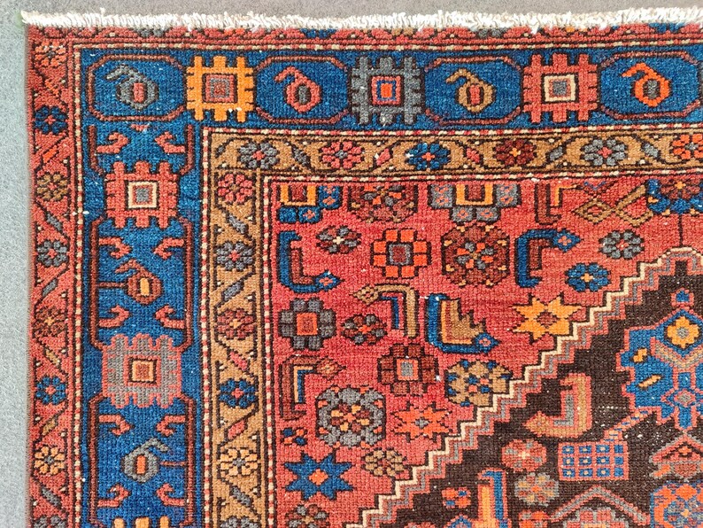 May include: Close-up of a section of a Persian rug with a detailed geometric pattern. The rug features a vibrant color palette of red, blue, orange, and brown. The border is a deep blue with repeating geometric shapes. The central field is red with intricate floral and geometric designs.