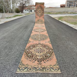 May include: A long, rectangular, vintage rug with an intricate, symmetrical pattern. The rug features a salmon-colored background with detailed designs in shades of brown, blue, and cream. The rug is displayed outdoors on a paved surface.