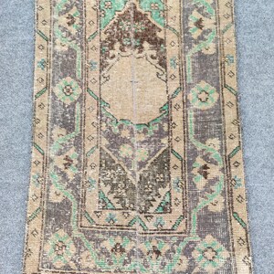 May include: A rectangular, distressed rug with a central beige panel and a dark gray border. The rug features a repeating floral pattern in shades of green, brown, and blue. The rug's design has a vintage, worn appearance.