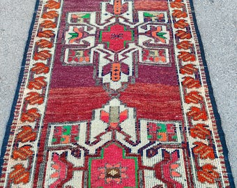 May include: A rectangular, hand-woven rug with a central geometric pattern in shades of red, pink, and cream. The rug features a border of orange leaf-like designs against a dark blue background. The rug's design is symmetrical and detailed.