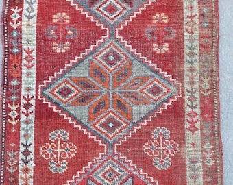 May include: Close-up of a red and grey patterned rug. The rug features geometric designs in shades of red, grey, orange, and white. The central design is a diamond shape with a starburst pattern. The rug's border has a repeating floral pattern.