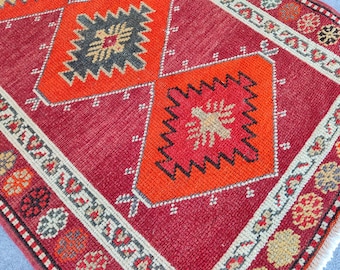 May include: A woven rug featuring a deep red background with large orange diamond shapes containing dark grey and beige geometric patterns. The border displays a repeating motif of small circular elements in orange, black, and beige tones.