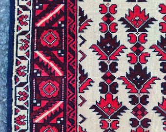 May include: Close-up of a beige and red patterned rug. The rug features a repeating floral design in red and dark red against a beige background. A dark blue border frames the rug.