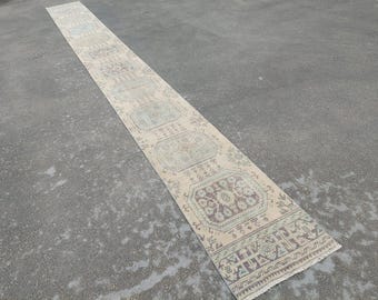 May include: A long, rectangular beige runner rug with a repeating geometric pattern in shades of brown, blue, and green. The rug has a fringed edge and is placed on a grey surface.