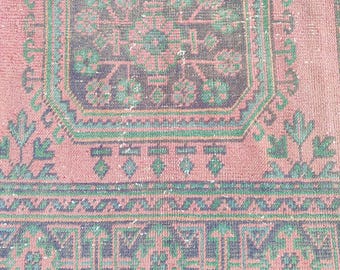 May include: A close-up of a vintage rug with a faded, distressed appearance. The rug features a central medallion design in shades of green and navy blue, set against a dusty rose background. Intricate geometric patterns and floral motifs are visible.