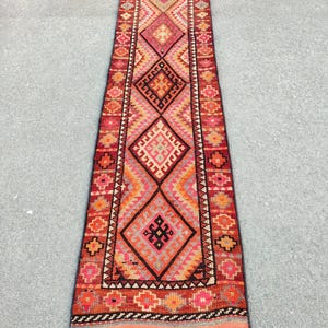 May include: A long, rectangular rug with a geometric pattern. The rug features a central design of large diamonds in shades of red, orange, and pink, bordered by smaller geometric shapes. The rug's edges are finished with a fringe.