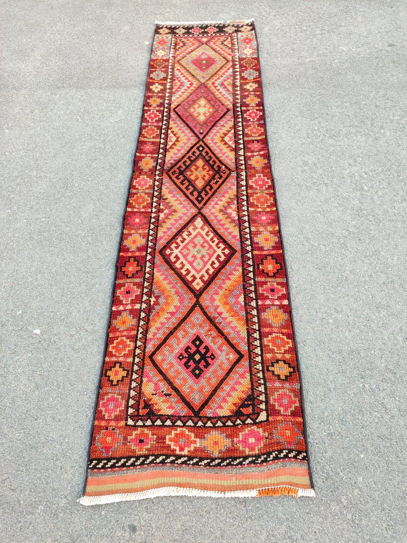 May include: A long, rectangular rug with a geometric pattern. The rug features a central design of large diamonds in shades of red, orange, and pink, bordered by smaller geometric shapes. The rug's edges are finished with a fringe.