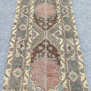 Pode incluir: Um tapete retangular de estilo vintage com um padrão central em tons de castanho, rosa e verde. O tapete apresenta um design geométrico repetitivo com detalhes florais e uma borda em tons suaves de bege e cinza. O tapete é feito de material tecido.
