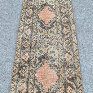 May include: A rectangular rug with a distressed appearance. It features a central diamond pattern in peach and black, framed by floral and geometric designs in green, brown, and beige. The rug has a vintage, worn look.