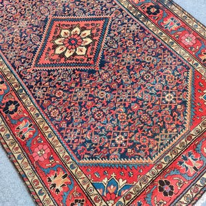 Peut inclure: Un tapis rectangulaire pr&eacute;sentant un champ central bleu fonc&eacute; avec un motif floral g&eacute;om&eacute;trique r&eacute;p&eacute;titif et un grand m&eacute;daillon en forme de losange. Le tapis est encadr&eacute; par une large bordure rouge orn&eacute;e de motifs floraux complexes en bleu et cr&egrave;me.