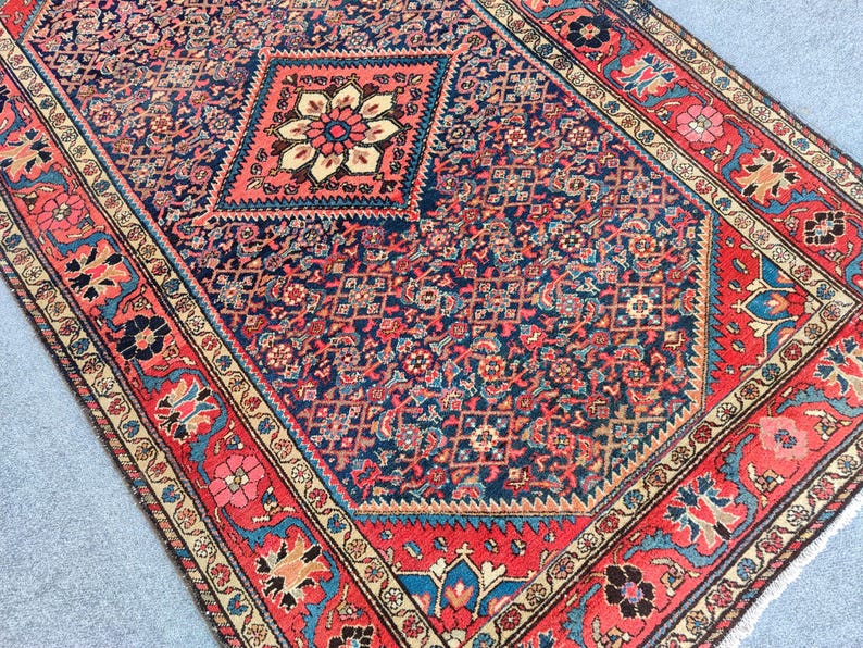 Peut inclure: Un tapis rectangulaire pr&eacute;sentant un champ central bleu fonc&eacute; avec un motif floral g&eacute;om&eacute;trique r&eacute;p&eacute;titif et un grand m&eacute;daillon en forme de losange. Le tapis est encadr&eacute; par une large bordure rouge orn&eacute;e de motifs floraux complexes en bleu et cr&egrave;me.
