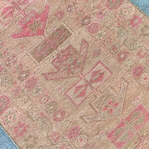 May include: Close-up of a vintage rug with a faded, distressed appearance. The rug features a repeating pattern of geometric shapes in shades of pink and brown against a beige background. The rug's texture is visible, suggesting a woven construction.