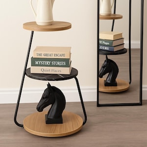 May include: A three-tiered side table with a black metal frame and wooden shelves. The top shelf holds a cream-colored teapot. The middle shelf displays three books. A black horse head sculpture sits on the bottom shelf. A tall black mirror is on the right.