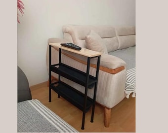 Puede incluir: Una mesa auxiliar de metal negro y madera con tres estantes. El estante superior es de color madera clara, y los estantes inferiores son negros. Un mando a distancia está sobre el estante superior. La mesa está junto a un sofá beige.