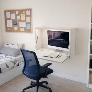 May include: A contemporary bedroom scene with a wall-mounted desk and computer, a blue mesh office chair, and a white bookcase filled with books. The room also includes a bed with geometric bedding and a corkboard with notes.