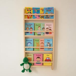May include: A light wooden bookshelf displaying a collection of children's books. The books feature titles such as "Coloring Book", "Bedroom", and "Car". A green soft toy turtle is placed on the bottom shelf. The books are in multiple languages.