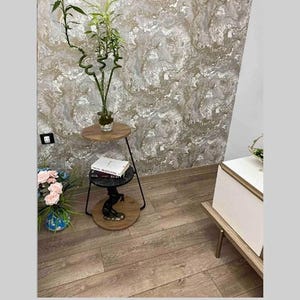 Può includere: Un tavolino da salotto a due livelli in metallo nero con un piano rotondo in legno. Il tavolino ha una scultura decorativa in metallo nero sul livello inferiore. Il tavolino è posizionato davanti a un muro con un motivo marmoreo.