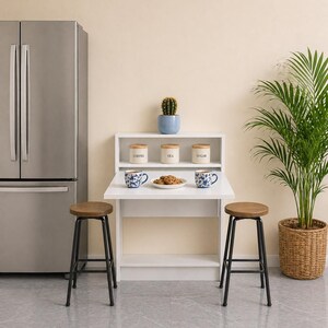 May include: A white, fold-down kitchen table with a shelf, two stools, and a stainless steel fridge. The table has two blue floral mugs and a plate of biscuits. A potted cactus and a plant in a woven basket are also present.