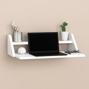 May include: A white wall-mounted desk with two shelves. A black laptop sits on the lower shelf, along with a notebook and glasses. The top shelf holds a small cactus in a white pot, a metal container with art supplies, and a white sphere.