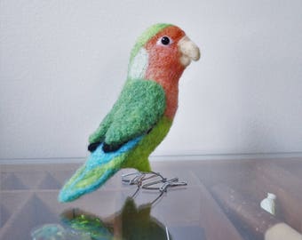 May include: A green and pink lovebird made of felt with a blue and green tail. The bird is perched on a wire stand.