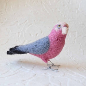 May include: A pink and grey felt bird with a black tail and wire legs. The bird is standing on a white surface.