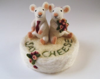 May include: Two white felt mice wearing a tie and a flower crown, sitting on a white felt cake with the words "Say Cheese" embroidered on it.