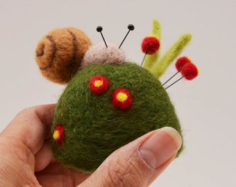 Puede incluir: Un cojín de alfileres de fieltro verde con forma de caracol con una concha de fieltro marrón, flores de fieltro rojo y amarillo y alfileres negros.