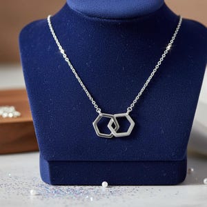 May include: A silver necklace with two interlocking hexagonal pendants. The necklace is displayed on a dark blue velvet jewelry display. The chain is delicate and the pendants are geometric.