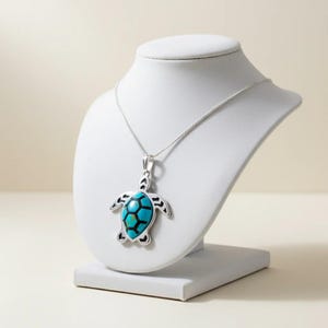 May include: A silver-toned turtle pendant necklace with a turquoise and black inlay. The turtle pendant is displayed on a white jewelry bust. The necklace chain is also silver-toned.