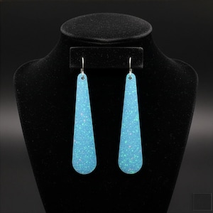 May include: A pair of light blue, teardrop-shaped earrings with a speckled, iridescent finish. The earrings are displayed on a black jewelry bust. The earrings have a simple, silver hook.