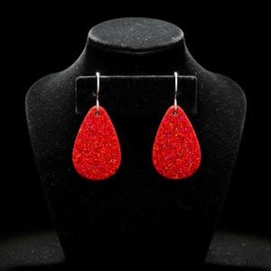May include: A pair of red teardrop earrings with a glittery, speckled appearance. The earrings hang from silver-colored hooks and are displayed on a black jewelry bust. The earrings are a vibrant red color and appear to be made of resin or a similar material.
