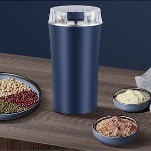 Pode incluir: Um moedor de especiarias elétrico azul marinho com tampa transparente, ao lado de pequenas tigelas de especiarias moídas e um prato de feijão. O moedor tem formato cilíndrico com ranhuras verticais.