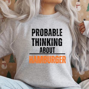Pode incluir: Um moletom cinza claro com o texto "PROBABLE THINKING ABOUT HAMBURGER" em preto e laranja. O moletom é de estilo casual, adequado para o uso diário.