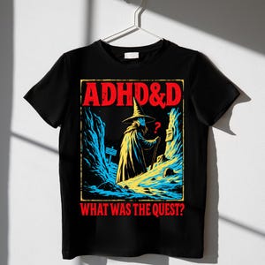 May include: A black t-shirt featuring a graphic print of a wizard in a pointed hat standing in a rocky landscape. The text at the top reads ADHD&D in red, and the text at the bottom reads WHAT WAS THE QUEST? in red. The wizard holds a staff.