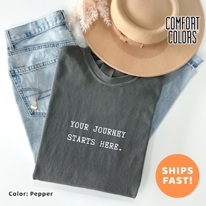 May include: A flat lay composition showing a pepper-coloured t-shirt with the text "YOUR JOURNEY STARTS HERE." The shirt is styled with light blue jeans, a tan hat, and a delicate necklace. The image also includes the text "COMFORT COLORS" and "SHIPS FAST!"