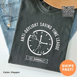 May include: Pepper-colored t-shirt with the text "ANTI-DAYLIGHT SAVING TIME LEAGUE" surrounding a cracked clock graphic. The shirt also features "EST. BIANNUALLY" below the clock. The shirt is folded on top of blue jeans.