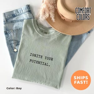 Może przedstawiać: Jasnozielona koszulka z napisem "IGNITE YOUR POTENTIAL." jest ułożona na niebieskich dżinsach. Widoczny jest również beżowy kapelusz i złoty naszyjnik. Koszulka jest marki "Comfort Colors", a kolor to "Bay".