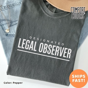 May include: A dark gray t-shirt with the words "DESIGNATED LEGAL OBSERVER" printed in white. The shirt is folded on top of a pair of blue jeans. The shirt is labeled "Comfort Colors" and the color is "Pepper".