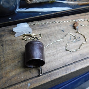 Könnte beinhalten: Ein rustikaler Glockenanhänger mit einer goldfarbenen Kette, verziert mit klaren Quarz Kristallen und einem kleinen geschnitzten Holz Akzent. Die Glocke ist aus dunklem, gealtertem Metall. Die Kette liegt auf einer Holzoberfläche.