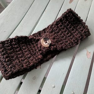 Op de afbeelding: Een donkerbruine, gehaakte hoofdband met een knoopsluiting. De hoofdband is gemaakt van dik garen en heeft een getextureerd uiterlijk. De knoop is lichtbruin en is op de voorkant van de hoofdband genaaid.