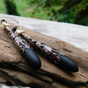 May include: A pair of black and copper dangle earrings with a filigree design. The earrings feature a black teardrop-shaped bead with a copper filigree design at the top.