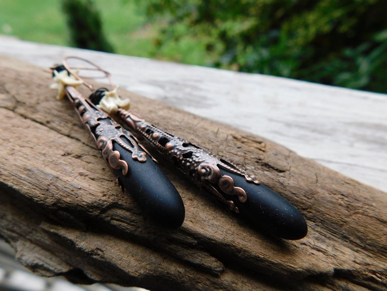 May include: A pair of black and copper dangle earrings with a filigree design. The earrings feature a black teardrop-shaped bead with a copper filigree design at the top.