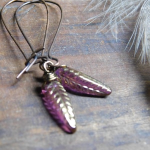 May include: Artisan earrings with purple, teardrop-shaped beads and gold detailing, suspended from dark metal ear wires. The earrings have a unique design. A feather is in the background. The beads are approximately 2 cm in length.