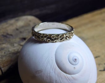 May include: A gold-toned ring with a textured, organic design. The ring is set against a white, spiral-shaped seashell, creating a contrast of textures and colors. The ring's surface is covered in small, raised details.