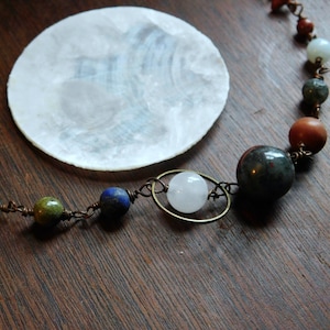 May include: A necklace featuring an assortment of colourful beads, including turquoise, green, blue, and white. The beads are strung on wire with a metal clasp. The necklace is displayed on a wooden surface with a white shell.