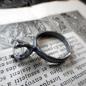 May include: A silver ring with a claw design holding a clear gemstone. The ring is on a vintage book with French text.