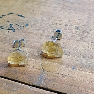 Puede incluir: Un par de pendientes de bot&oacute;n con piedras preciosas de citrino amarillo dorado en bruto. Los pendientes tienen postes y cierres plateados. Las piedras preciosas tienen una textura rugosa y natural, sobre una superficie de madera.