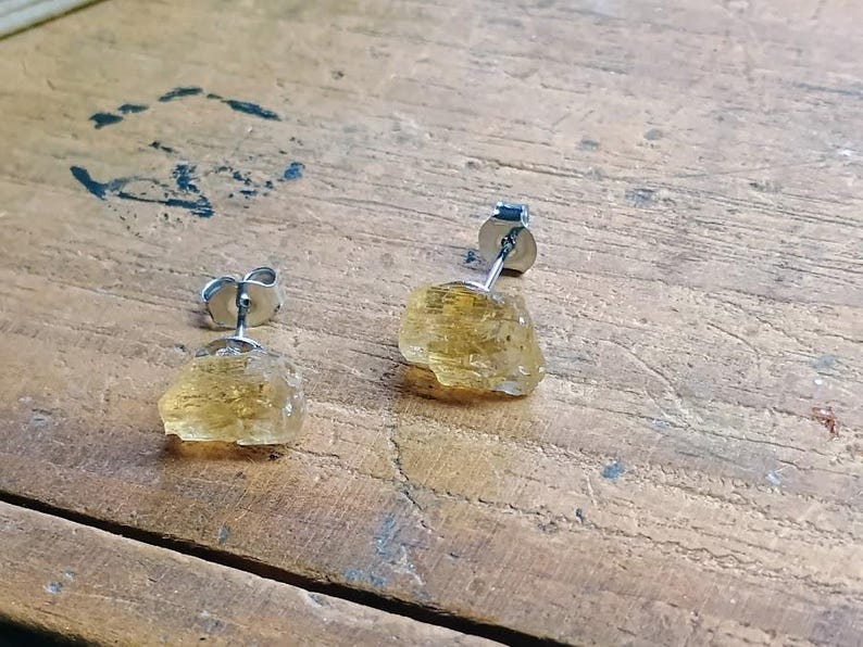 Puede incluir: Un par de pendientes de bot&oacute;n con piedras preciosas de citrino amarillo dorado en bruto. Los pendientes tienen postes y cierres plateados. Las piedras preciosas tienen una textura rugosa y natural, sobre una superficie de madera.